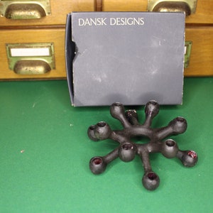 Könnte beinhalten: Ein schwarzer Kerzenhalter aus Metall mit einem Sternendesign und neun Kerzenhaltern. Der Kerzenhalter steht auf einer grünen Oberfläche. Die Schachtel trägt die Aufschrift "DANSK DESIGNS".
