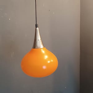 Op de afbeelding: Oranje hanglamp met een chromen bovenkant en een druppelvormige glazen kap. De lamp hangt aan een zwart snoer en een chromen plafondbevestiging. Het licht heeft een levendige oranje kleur.