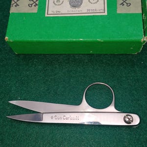 Pode incluir: Tesoura prateada com um grande orifício para o dedo e o texto "Don Carlos" na lâmina. A tesoura é exibida sobre uma superfície verde, com uma caixa verde ao fundo.