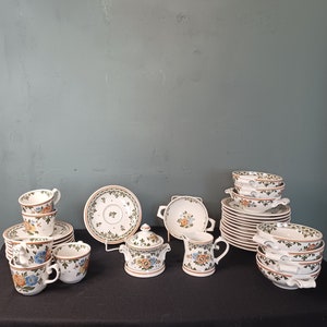 Puede incluir: Un juego de vajilla de cerámica blanca con un patrón floral en tonos verdes, naranjas y azules. El juego incluye tazas, platillos, tazones, un azucarero y una jarra de crema.