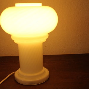 Könnte beinhalten: Eine weiße Tischlampe mit einem runden, mattierten Glasschirm. Die Lampe ist eingeschaltet und das Licht beleuchtet den Schirm.