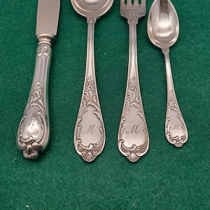 Könnte beinhalten: Ein Satz antiker Silberbestecke, darunter ein Messer, ein Löffel, eine Gabel und ein kleinerer Löffel, alle mit verzierten Griffen und dem Buchstaben "M" graviert. Das Besteck ist silberfarben und liegt auf einer grünen Oberfläche.