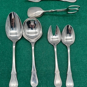 May include: A selection of antique silver serving cutlery, including spoons, forks, and a serving fork. The silverware is arranged on a green surface. The utensils have ornate handles and a polished finish.