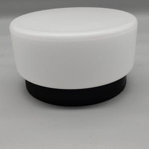 Può includere: Una copertura cilindrica per lampada caratterizzata da una parte superiore in vetro bianco opaco liscio e una base circolare nera solida. L'oggetto presenta un profilo pulito con una superficie superiore piatta e lati verticali per il montaggio.