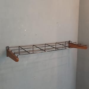 Puede incluir: Estante de pared con un diseño de rejilla de alambre negro y soportes de madera. El estante es rectangular y parece estar hecho de metal y madera. El estante está montado en una pared gris.