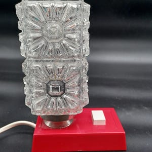 May include: A vintage table lamp with a clear, textured glass shade featuring a geometric design. The lamp sits on a red base with a white on/off switch. The lamp's dimensions are approximately 10 cm wide and 20 cm tall.