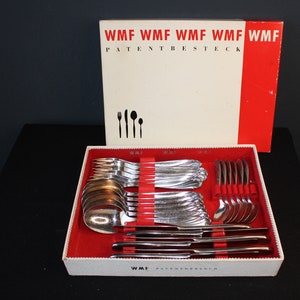 Op de afbeelding: Een vintage WMF bestekset in een rood-witte doos. De doos heeft de tekst "WMF PATENTBESTECK" op de bovenkant. Het bestek is in rijen in de doos gerangschikt, met de vorken, messen en lepels gescheiden.