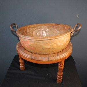 Puede incluir: Un cuenco de cobre antiguo grande con dos asas de hierro. El cuenco está sentado en un soporte de madera con tres patas.