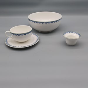 Puede incluir: Un juego de té de porcelana blanca con ribete decorativo azul y verde. El juego incluye una taza y un platillo, un cuenco pequeño y un cuenco para servir más grande. El ribete azul presenta un patrón repetitivo. El juego se muestra sobre un fondo gris.
