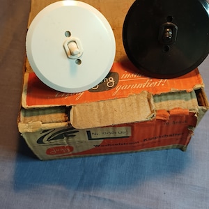 Puede incluir: Dos placas de interruptor de luz, una blanca y una negra, se muestran en la parte superior de una caja de cartón. La caja está etiquetada como "Jung" y "Nr. 10/501 Ub" y tiene un diseño rojo y blanco. Las placas de interruptor son redondas y tienen un solo interruptor de palanca.