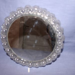 May include: A round mirror with a clear, textured frame made of small, round beads. The mirror reflects a room with a white door.