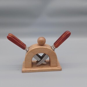 Può includere: Un set di coltelli da formaggio in legno con finitura naturale. Il set include un supporto in legno con un design curvo, una piccola palla di legno sulla parte superiore e due coltelli da formaggio con manici rossi e dettagli argentati. I coltelli sono lunghi circa 15 cm.