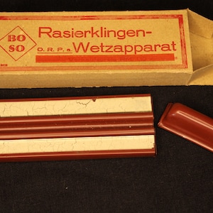 May include: A vintage razor blade sharpener in a cardboard box. The box is labeled "Rasierklingen-Wetzapparat" and "BoSo". The sharpener is made of metal and has a red and white finish.