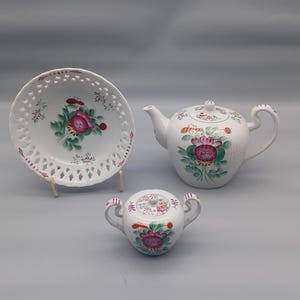 May include: A white porcelain tea set comprising a teapot, sugar bowl, and a plate. Each item is adorned with a floral pattern in pink, red, and green. The plate has a pierced edge.