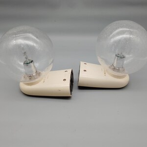 May include: A pair of wall-mounted light fittings featuring off-white, rounded plastic bases and clear, spherical glass globes with a subtle speckled texture. Each fitting has a metallic bulb socket visible inside the glass sphere.