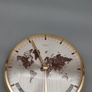 May include: A round Kienzle Quartz world map clock with a gold-tone metal frame and base. The clock face features a brown world map graphic, thin gold hands, and a curved 24-hour scale at the bottom with black numerals ranging from 1 to 24.