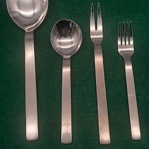 May include: A collection of four stainless steel cutlery items: a large spoon, a small spoon, and two forks. The utensils feature a minimalist design with long, flat handles and rounded spoon bowls. The forks have three and four prongs.