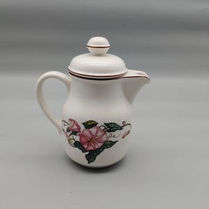 Może przedstawiać: Biała ceramiczna śmietanka z pokrywką i uchwytem. Śmietanka jest ozdobiona różowymi kwiatami powoju i zielonymi liśćmi. Obręcz i pokrywka są wykończone brązowym paskiem.