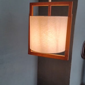 Könnte beinhalten: Eine Hängelampe mit Holzrahmen und einem weißen Stoffschirm, der an einem weißen Kabel hängt. Die Lampe ist eingeschaltet und strahlt ein warmes Licht aus.