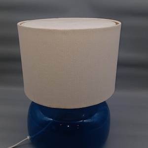 Könnte beinhalten: Eine blaue Keramik-Tischlampe mit cremefarbenem Stoffschirm. Die Lampe hat einen runden Sockel und einen zylindrischen Schirm. Die Lampe ist mit einem weißen Kabel angeschlossen.