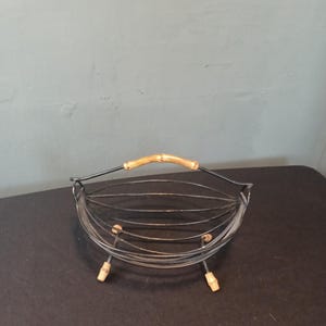 Op de afbeelding: Een zwarte draadmand met een houten handvat en houten poten. De mand is ontworpen om fruit of andere items in te bewaren.