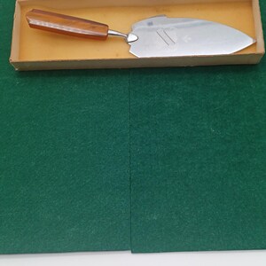 Könnte beinhalten: Ein Vintage-Kuchenheber mit einer Edelstahlklinge und einem braunen Griff, präsentiert in einer Pappschachtel mit rotem Deckel. Der Heber ist zum Schneiden und Servieren von Kuchen und Gebäck bestimmt.