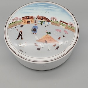 以下が含まれることがあります： 蓋付きの丸い白い陶器製の箱で、蓋には農場の風景がカラフルに描かれています。絵には、熊手を持った人物、バケツを運ぶ人物、農場の建物、豚、鶏が描かれています。LAPLAU 3という文字が印刷されています。