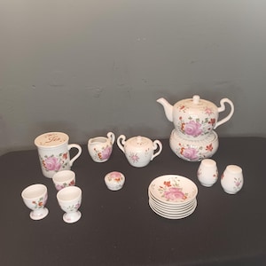 Peut inclure: Un service à thé en porcelaine blanche avec des motifs floraux roses et rouges. L'ensemble comprend une théière, un pot à crème, un sucrier, deux tasses, deux coquetiers, deux salières et poivrières et une pile de soucoupes.