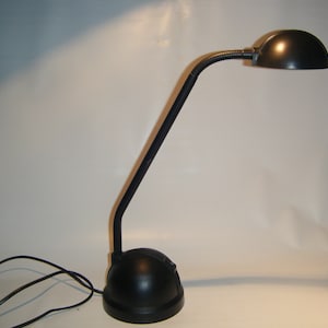 Könnte beinhalten: Eine schwarze Schreibtischlampe mit einem flexiblen Schwanenhalsarm und einem runden Schirm. Die Lampe hat einen schwarzen Sockel und ein schwarzes Kabel.