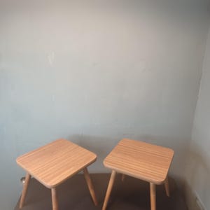 Puede incluir: Dos mesas auxiliares cuadradas de madera con un acabado de madera de grano marrón claro y cuatro patas delgadas.