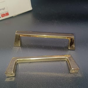 Peut inclure: Deux poignées de meuble vintage en laiton. Chaque poignée a une barre rectangulaire et des supports coudés. Les poignées sont de couleur or chaud et semblent neuves. La quincaillerie est probablement pour un tiroir ou une porte d'armoire.