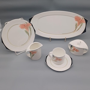 May include: A white porcelain dinner service with a floral pattern. The set includes a large oval serving dish, a round plate, a creamer jug, a cup and saucer, and a sugar bowl. Each piece has a black border and a pink and green floral design.