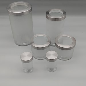 Può includere: Un set di sei barattoli cilindrici in vetro con coperchi in metallo argento spazzolato. I contenitori presentano una finitura in vetro smerigliato sulle sezioni inferiori e cime in vetro trasparente, disposti in altezze variabili.