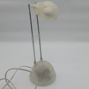 Può includere: Una piccola lampada da scrivania bianca con base arrotondata e braccio flessibile e regolabile. La testa della lampada è anch'essa arrotondata e traslucida. La lampada ha un braccio metallico color argento e un cavo di alimentazione bianco.