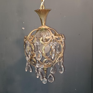 Könnte beinhalten: Verzierter goldfarbener Kronleuchter mit klaren Kristallakzenten. Die Leuchte hat ein dekoratives filigranes Design um eine zentrale Lichtquelle, mit tropfenförmigen Kristallen, die darunter hängen. Eine Kette hängt den Kronleuchter auf.