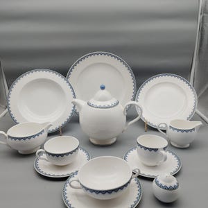 Puede incluir: Un juego de té de porcelana blanca con un borde con dibujos azules. El juego incluye una tetera, platos, tazas, platillos, una jarra de crema y un azucarero. El juego de té está dispuesto sobre una superficie gris.