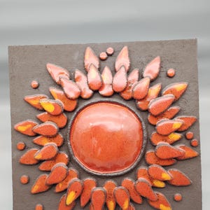 Könnte beinhalten: Ein quadratisches Wandkunstwerk aus Keramik. Es zeigt einen zentralen orangefarbenen Kreis, der von stilisierten orangefarbenen und gelben blütenblattartigen Formen umgeben ist. Der Hintergrund ist eine dunkelgraue, strukturierte Oberfläche. Das Stück hat ein glänzendes Finish.