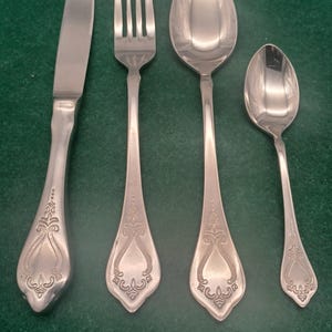 Può includere: Un set di quattro posate argentate: un coltello, una forchetta e due cucchiai. Ogni pezzo presenta un motivo floreale decorativo sul manico. Le posate sono disposte su una superficie verde.