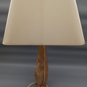 Op de afbeelding: Een tafellamp met een beige lampenkap en een bruine, patroonvoet. De lamp heeft een rechthoekige lampenkap en een snoer met een aan/uit-schakelaar. De voet heeft een unieke uitstraling van natuursteen.