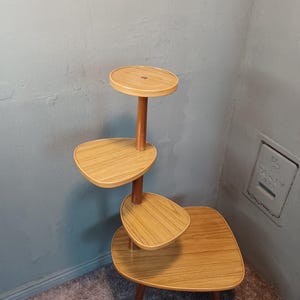 Puede incluir: Un soporte para plantas de estilo moderno de mediados de siglo con tres niveles. El soporte está hecho de madera y tiene un acabado marrón claro. El nivel superior es redondo y los dos niveles inferiores son ovalados.