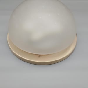 Op de afbeelding: Een koepelvormige lamp met een matwitte glazen kap met een subtiele gespikkelde textuur. De kap is gemonteerd op een ronde, lichtgekleurde metalen voet met een gladde, matte afwerking, wat zorgt voor een strak en eenvoudig ontwerp.