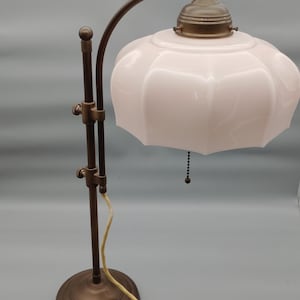 Puede incluir: Una lámpara de mesa con base de metal en tono bronce y brazo ajustable. La lámpara cuenta con una pantalla de vidrio festoneada de color rosa pálido y un interruptor de cadena. Un cable de alimentación claro sale de la base de la lámpara.