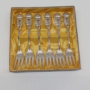 Pode incluir: Um conjunto de seis garfos prateados com desenhos florais, apresentados numa caixa forrada a cetim dourado. Os garfos têm detalhes intrincados de hastes e dentes. A caixa é retangular com uma borda decorativa.