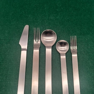 Può includere: Un set di posate in acciaio inossidabile composto da un coltello, due forchette e due cucchiai. Gli utensili sono disposti su una superficie verde scuro. Il coltello ha la punta arrotondata e i cucchiai sono di diverse dimensioni.