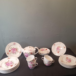 Könnte beinhalten: Ein Vintage-Teeservice mit Teekanne, Tassen, Untertassen und Tellern. Das weiße Porzellanset ist mit einem rosa Rosenblütenmuster und roten Akzenten verziert. Die Teller haben einen Durchmesser von ca. 23 cm.
