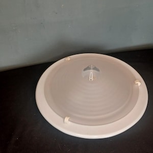 May include: White ceiling light fitting with a circular, frosted glass shade and a central, cone-shaped diffuser. The light fitting has three white plastic clips on the edge of the shade.