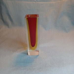 Puede incluir: Un jarrón de vidrio transparente con un interior rojo y amarillo. El jarrón tiene forma cuadrada y una abertura rectangular.