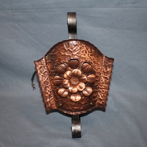 Op de afbeelding: Een koperen wandlamp met een gehamerde afwerking en een bloemmotief. De lamp heeft een zwarte metalen haak om op te hangen.