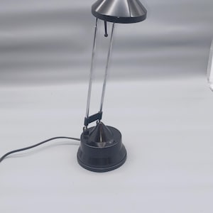 Può includere: Una moderna lampada da scrivania con paralume conico in metallo spazzolato e base nera. La lampada è dotata di un braccio cromato e di un cavo di alimentazione. La lampada è alta circa 30 cm.