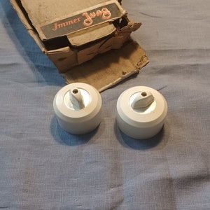 May include: Two white light switch covers with silver toggles. The covers are on a blue fabric surface. The box they came in is visible in the background. The box is brown cardboard with the text "Immer Jung" printed on it.